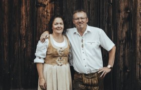 Wirtsleute Silvia und Franz Schrammel, © Niederösterreich Werbung/David Schreiber Ein Mann und eine Frau in traditioneller Tracht vor einer Holzwand.