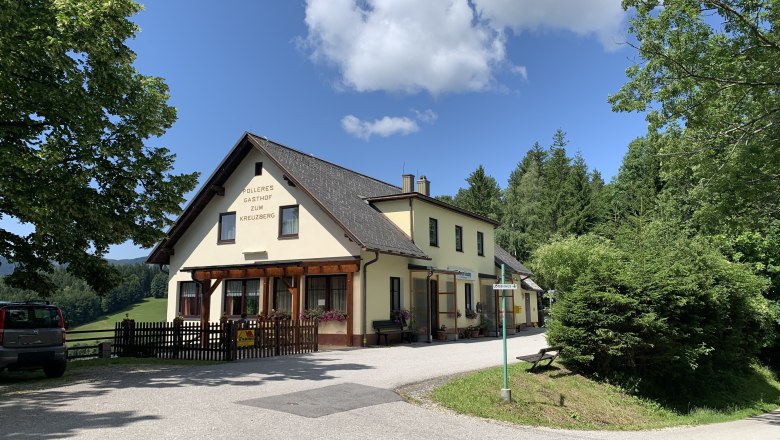 Gasthaus Polleres, © TVB Semmering-Rax-Schneeberg Ein gelbes Gasthaus mit der Aufschrift 'Polleres Gasthof zum Kreuzberg' umgeben von Bäumen und einem blauen Himmel.