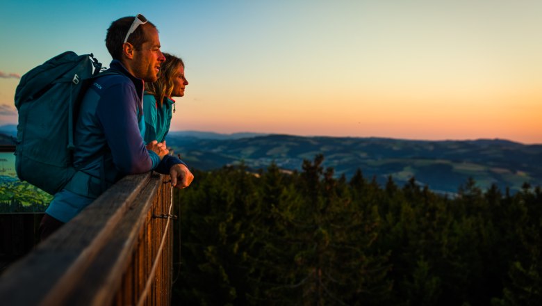 Sonnenaufgang auf der Aussichtswarte am Hutwisch, © Wiener Alpen/Martin Fülöp Zwei Personen stehen auf einem Aussichtspunkt und blicken in die Ferne bei Sonnenuntergang.