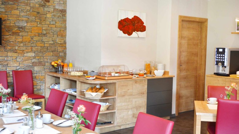 Frühstücksbuffet, © Familie Martin Ein Frühstücksbuffet in einem gemütlichen Raum mit roten Stühlen, Holzmöbeln und einer Steinwand. Auf dem Buffet sind Croissants, Müsli und Orangensaft.