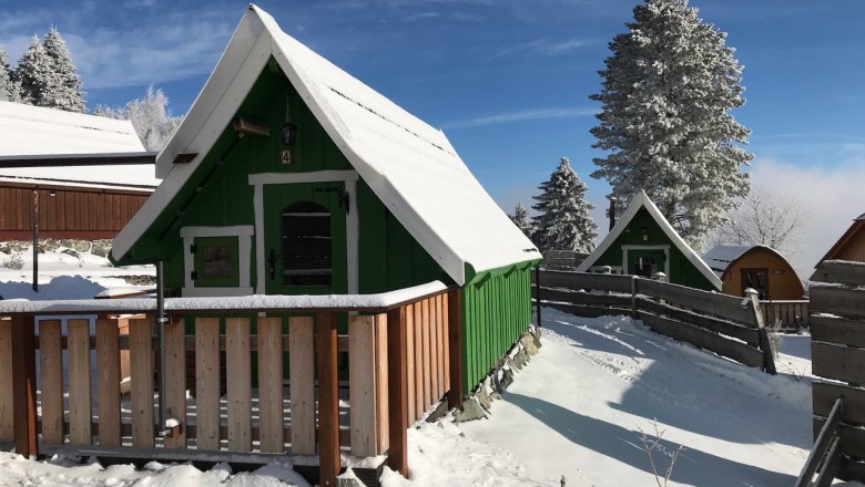 Wood Lodge, © Glamping-Park Mönichkirchen OG Verschneite Holzhütten in einer winterlichen Landschaft mit blauem Himmel.