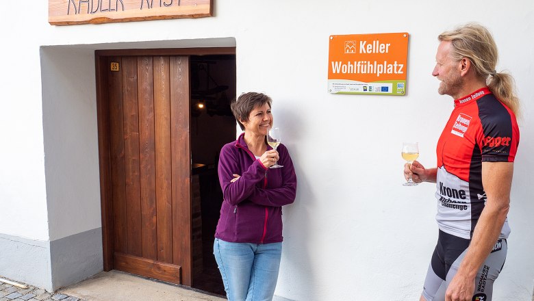 Keller-Wohlfühlplatz Jetzelsdorf, © Initiative Pulkautal / Mödl Zwei Personen stehen vor einem Gebäude mit der Aufschrift 'Radler-Rast' und 'Keller Wohlfühlplatz', sie halten Weingläser in der Hand.