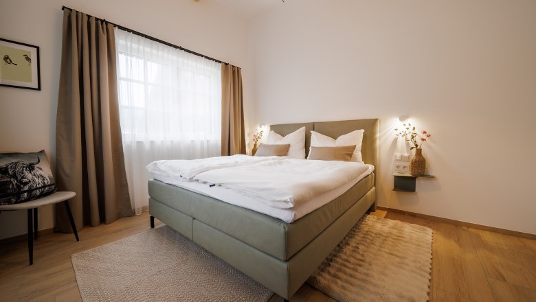 Schlafzimmer, © Göllitzhof Modernes Schlafzimmer mit Doppelbett, Holzboden und großen Fenstern mit Vorhängen.