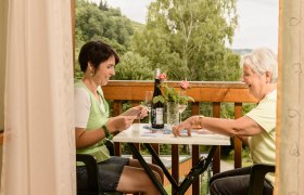 Frühstück draussen, © Rita Newman Zwei Frauen spielen Karten auf einem Balkon mit Wald im Hintergrund.