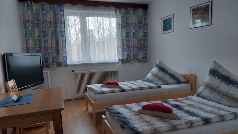 TDR-Pension Welserhof, © Bartfai Ein einfach eingerichtetes Zimmer mit zwei Einzelbetten, einem Tisch, einem Fernseher und einem Fenster mit bunten Vorhängen.