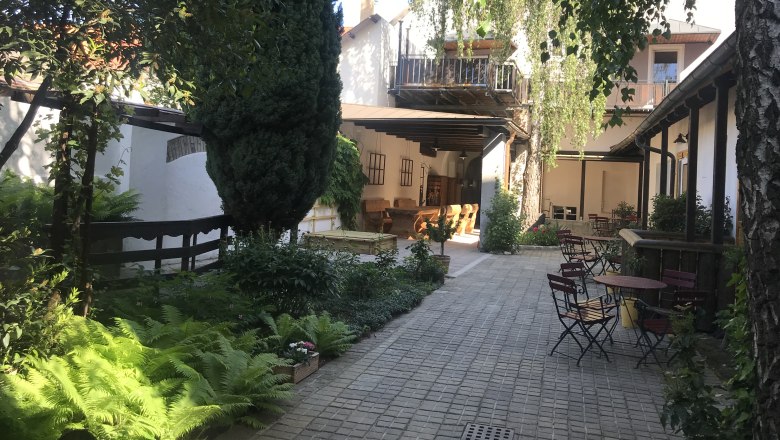 Innenhof, © Fam. Lahner Ein ruhiger Innenhof mit Bäumen, Pflanzen und Gartenmöbeln unter blauem Himmel.