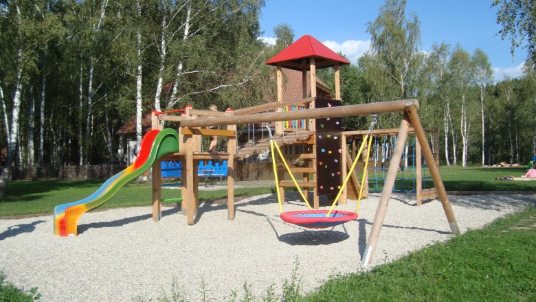 Kinderspielplatz, © Hannes Messmann Ein Kinderspielplatz mit Rutsche, Schaukel und Klettergerüst, umgeben von Bäumen.