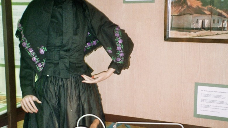 Trachtenpuppe, © Adolf Haider Eine Schaufensterpuppe in traditioneller Kleidung neben einem alten Kinderwagen in einem Ausstellungsraum.
