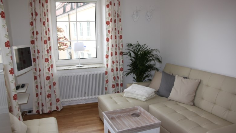 Altes Pförtnerhaus, © © Elisabeth Hofer Helles Wohnzimmer mit beigem Sofa, Kissen, Couchtisch und Fenster mit Vorhängen.