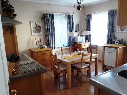 Küche, © bed and pool Gemütliche Küche mit Holzmöbeln, Esstisch und Stühlen, zwei Fenstern mit Vorhängen, und Küchenutensilien auf der Arbeitsfläche.