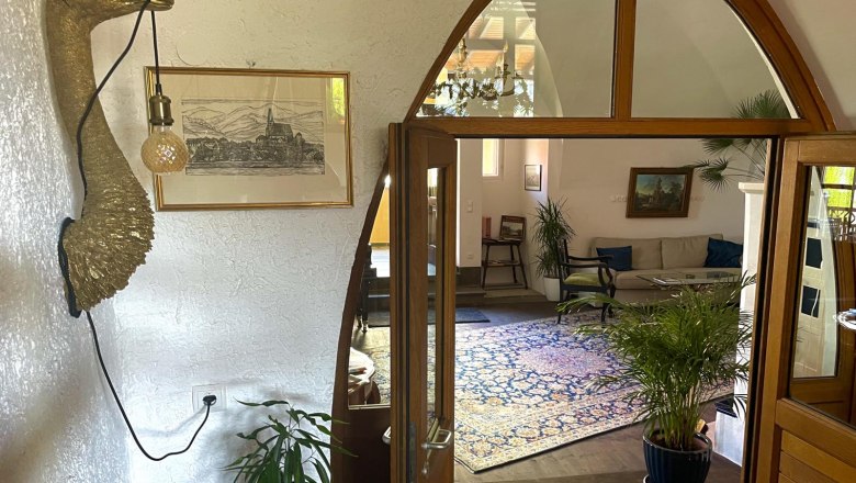 Abgang, © Georg Smolka Innenansicht eines Wohnzimmers mit Pflanzen, einem Sofa und einem dekorativen Wandobjekt in Form eines Vogels mit einer Lampe.