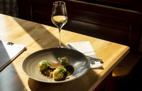 Geschmorter Schweinebauch, Chilikraut, Erdäpfelknödel in knusperiger Schweineschwarte gewälzt, © Niederösterreich Werbung/Michael Reidinger Teller mit Fleisch, Knödeln und Soße, Weinglas auf Holztisch im Sonnenlicht.