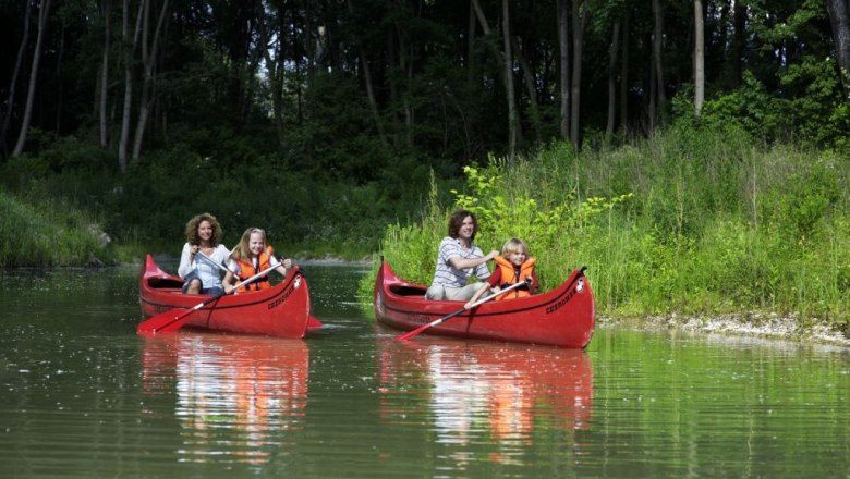 Wasserpark Tulln, © DIE GARTEN TULLN Zwei rote Kanus mit Erwachsenen und Kindern auf einem ruhigen Fluss, umgeben von grüner Vegetation.