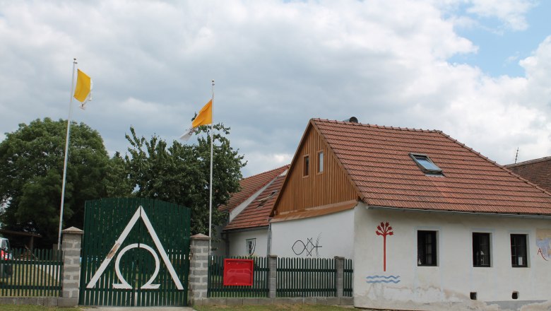 Haus Agnes 3, © Hannes Mesmann Ein Haus mit roten Dachziegeln, grüner Zaun mit Alpha-Omega-Symbol, zwei gelbe Fahnen und Wandmalereien.