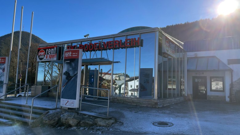 Sport-Puschi-Verleih-Bergbahn, © Sport Puschi Außenansicht eines Skiverleihs mit Glasfassade und Werbeschildern, umgeben von Bergen und strahlendem Sonnenschein.