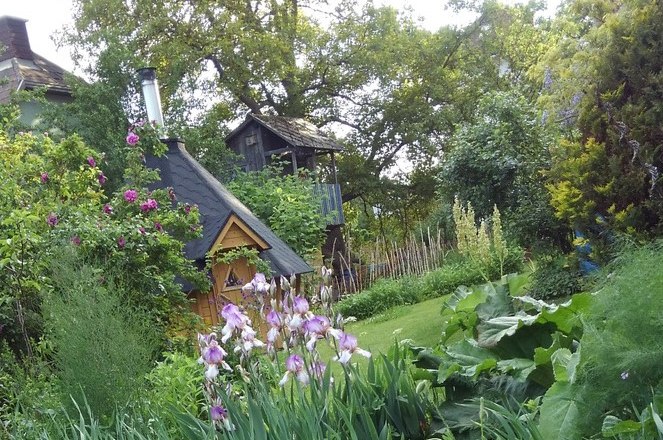 finnhaus-bluetenwald3_ausschn_c_edith-weiss, © Edith Weiß Ein idyllischer Garten mit einem kleinen Holzhaus, umgeben von blühenden Pflanzen und Bäumen.