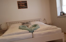 Zimmer, © mittendrin Dorfpension zum Ehn Weissesdoppelbett mit einer Decke, welche ein Herz formt. Über dem Bett hängt auf einer weißen Wand ein rotes Bild.