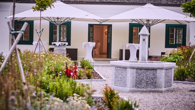 Brunnen im Außenbereich, © Franz Gleiß Ein steinerner Brunnen in einem Garten vor einem Gebäude mit weißen Sonnenschirmen und grünen Fensterläden.