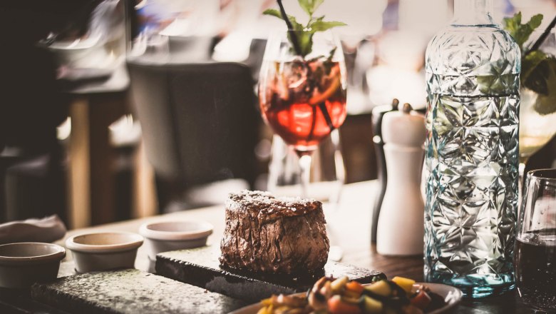 Steak nach Schweizer Art, © Route 66 Fehr Ein saftiges Steak auf einem heißen Stein serviert, begleitet von Gemüse, einer Karaffe Wasser und einem Cocktailglas.