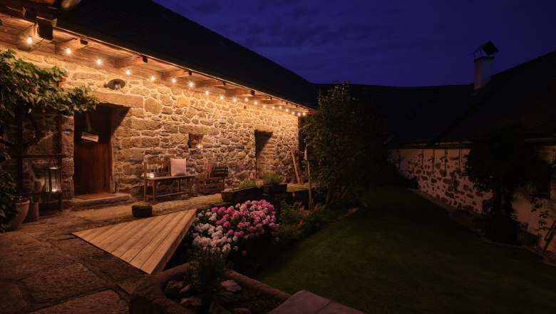 Aigen13, © Mathias Lixl, lixl.com Ein beleuchteter Innenhof bei Nacht mit Steinmauern, Holzbänken und blühenden Blumen.