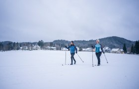 Langlaufen in Karlstift, © Waldviertel Tourismus, Robert Herbst Zwei Personen beim Langlaufen auf einer schneebedeckten Fläche vor einem Dorf und bewaldeten Hügeln.