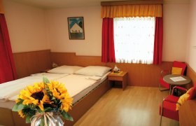 Zimmerfoto, © Landgasthof Oppolzer Ein gemütliches Zimmer mit Doppelbett, roten Vorhängen, einem Tisch mit Stühlen und einem Sonnenblumenstrauß im Vordergrund.