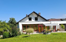 Hausansicht, © Ferienwohnung Penz Stolzenthal, Fotograf Hermine Penz Ein weißes Haus mit rotem Rahmen und Garten vor blauem Himmel.
