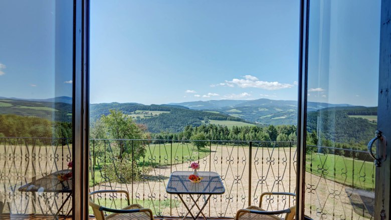 Ausblick vom Zimmer, © Wachahof Blick von einem Balkon auf eine hügelige Landschaft mit Wäldern und Feldern, zwei Stühle und ein Tisch mit Blumen im Vordergrund.