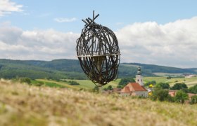 kunst_in_der_natur-002125_ausschn__c_natur_im_garten_alexander_haiden, © Natur im Garten/Alexander Haiden Kunstinstallation aus Metall in einer ländlichen Landschaft mit Kirche im Hintergrund.
