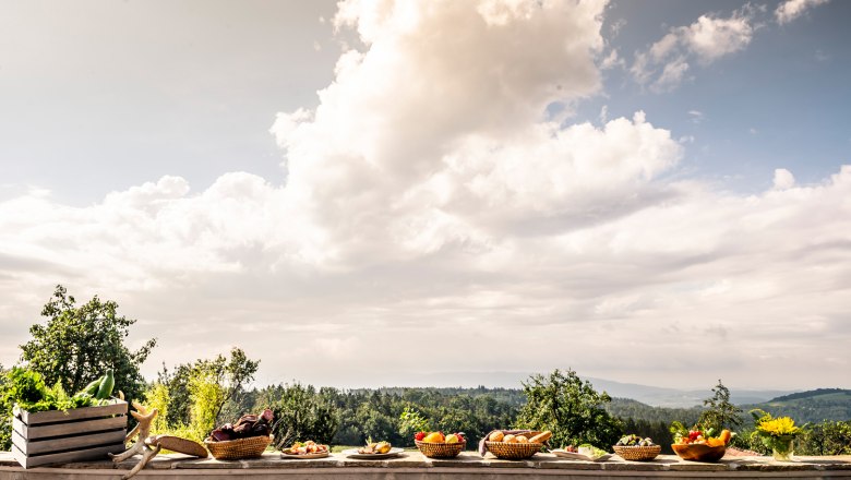 Reiche Auswahl an selbst erzeugten Produkten, © Robert Herbst Verschiedene Körbe mit Obst und Gemüse auf einer Mauer vor einer Landschaft mit Bäumen und Wolken.