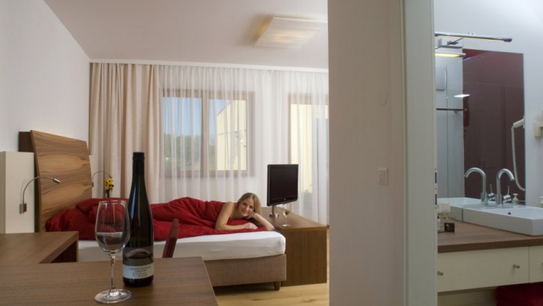 Kitzer Wein Genuss Zimmer, © Johannes Kitzler Ein modernes Hotelzimmer mit einem Bett, auf dem eine Person liegt, und einem Tisch mit einer Weinflasche und einem Glas.