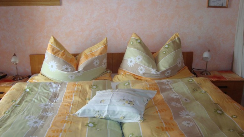 Schlafzimmer, © Fam. Sommer-Leitner Ein gemütliches Schlafzimmer mit einem Doppelbett, das mit gelb-grüner Bettwäsche und Blumendekor bezogen ist. Zwei Nachttischlampen stehen daneben.
