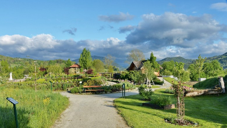 Paradiesgart’l Molzbachhof, © Hotel Molzbachhof Ein gepflegter Garten mit Wegen, Bäumen und Blumen, umgeben von grünen Hügeln und einem blauen Himmel mit Wolken.