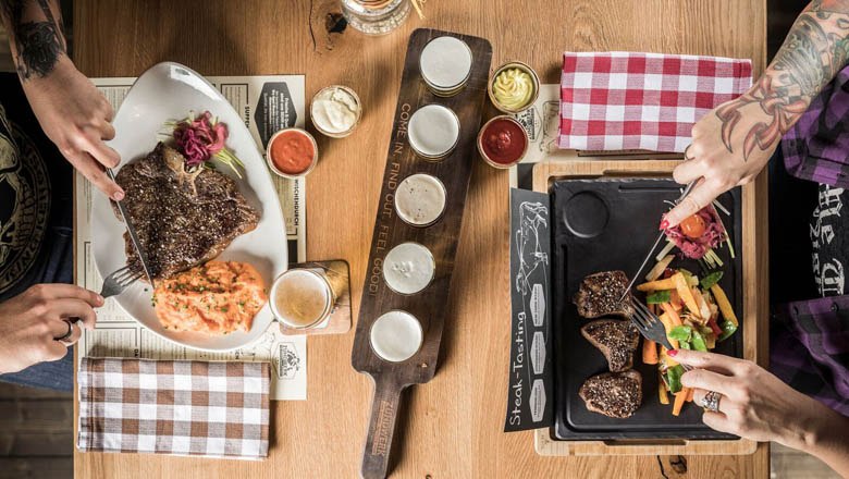 Zündwerk, © Lukas Kirchgasser Photography Zwei Personen essen Steaks mit Beilagen an einem Holztisch.
