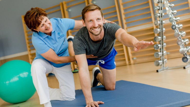 Bewegungstherapie im Gesundheitsresort Königsberg, © Niederösterreich Werbung/Doris Schwarz-König Ein Mann macht eine Übung auf einer Matte, unterstützt von einer Frau in einem Fitnessstudio.