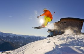 Action & Fun auf der Gemeindealpe-Terzerhaus, © Leiminger Ein Skifahrer springt vor dem Terzerhaus auf der Gemeindealpe in die Luft, umgeben von schneebedeckten Bergen und blauem Himmel.