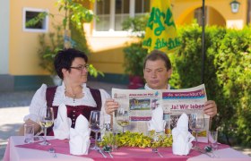 Gasthof Franzl, © Michael Rzepa Ein Mann und eine Frau sitzen an einem Tisch im Freien, der mit Gläsern und Besteck gedeckt ist. Der Mann liest eine Zeitung.