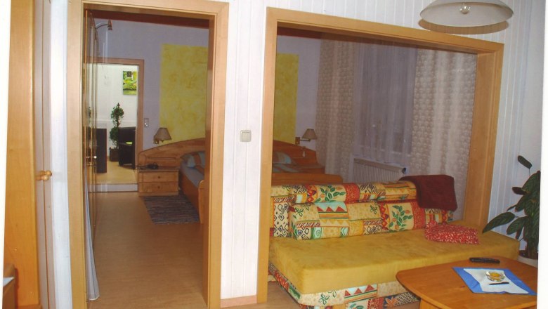 Gästezimmer Weixelbaum, © Weixelbaum Ein gemütliches Gästezimmer mit buntem Sofa, Holztisch und Blick auf ein Schlafzimmer mit Doppelbett.