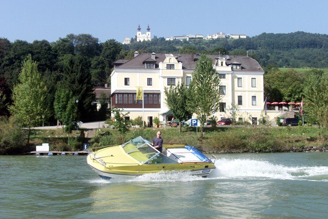 HausmitBoot, © Wachauerhof Renner GMBH Ein gelbes Motorboot fährt auf einem Fluss vor einem großen, hellen Gebäude mit Hügeln im Hintergrund.
