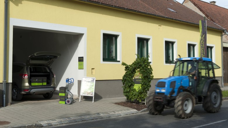 Weinauslieferung, © Weingut Holzmann Gelbes Gebäude mit geöffneter Garage, Auto und Traktor davor.