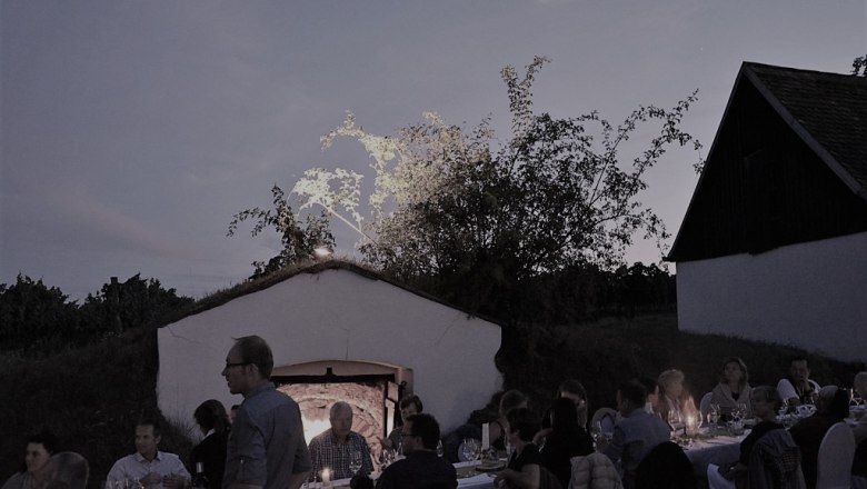 Tafeln in der Öhlbergkellergasse, © Weingut A+A Mayer Menschen sitzen an einem langen Tisch im Freien bei Dämmerung, umgeben von Gebäuden und Bäumen.