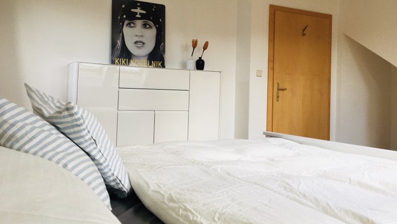 Schlafzimmer, © Foto: Eva Zwirner Modernes Schlafzimmer mit weißem Bett, gestreiften Kissen und Kommode. An der Wand ein Bild von Kiki Kogelnik und zwei Vasen mit Blumen.