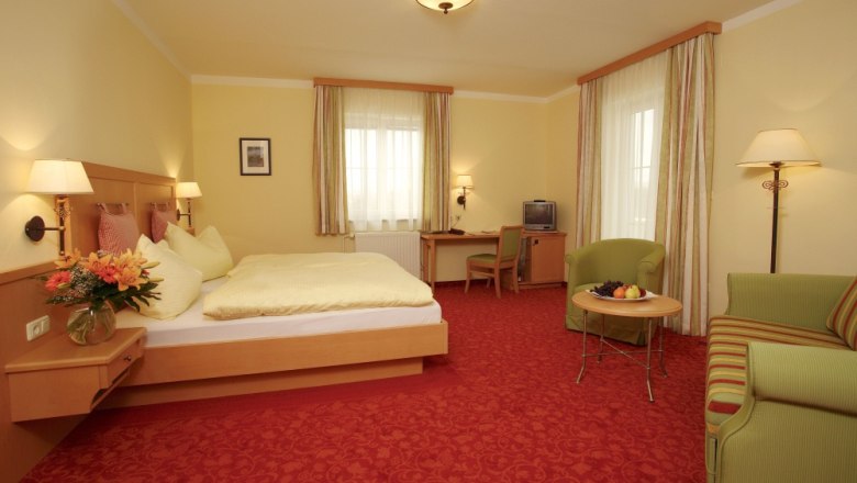 Doppelzimmer, © Hotel Wachau Ein gemütliches Hotelzimmer mit Doppelbett, Sofa, Tisch und Fernseher. Gelbe Wände und rote Teppiche dominieren das Zimmer.