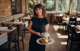 Wirtin Zanetá Haselberger, © Niederösterreich Werbung/Daniela Führer Frau serviert Essen in einem gemütlichen Restaurant mit Holzmöbeln.