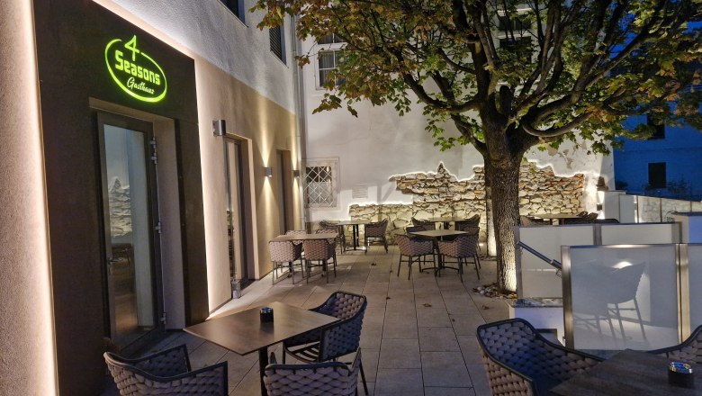 TD-Terasse 4 Season, © Hendler Außenterrasse eines Restaurants mit Tischen und Stühlen, beleuchtet bei Dämmerung, Baum im Hintergrund.