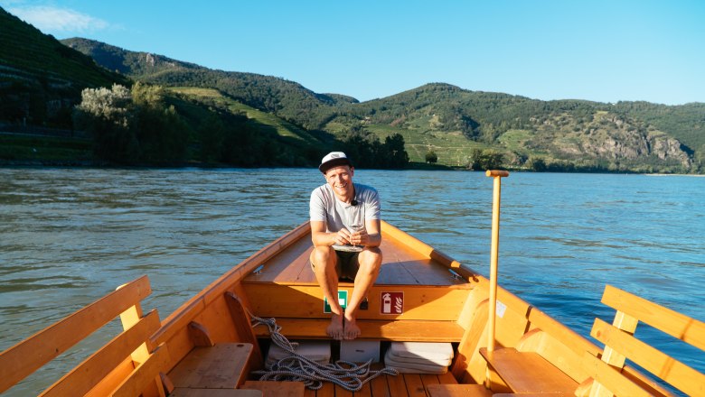 ahoi-wachau-steuerboard, © NÖW_Daniel Gollner Person sitzt auf einem Holzboot auf einem Fluss, umgeben von grünen Hügeln.