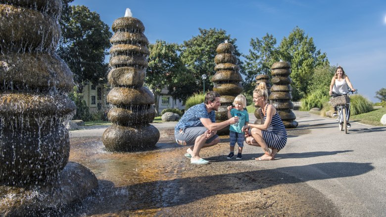 brunnen_c_robert-herbst-stadtgemeinde-tulln, © Robert Herbst/Stadtgemeinde Tulln Familie am Brunnen in einem Park, mit einer Frau auf einem Fahrrad im Hintergrund.
