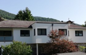 Außenansicht Ferienhaus Johanna, © Familie Schoßmann Außenansicht eines modernen weißen Ferienhauses mit Bäumen im Vordergrund und bewaldeten Hügeln im Hintergrund.