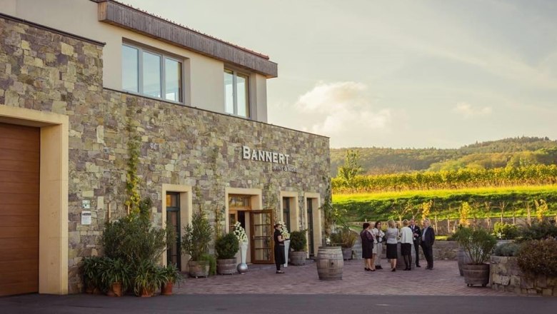 Weingut Bannert, © Weingut Bannert Weingut Bannert mit Menschen vor dem Gebäude und Weinbergen im Hintergrund.