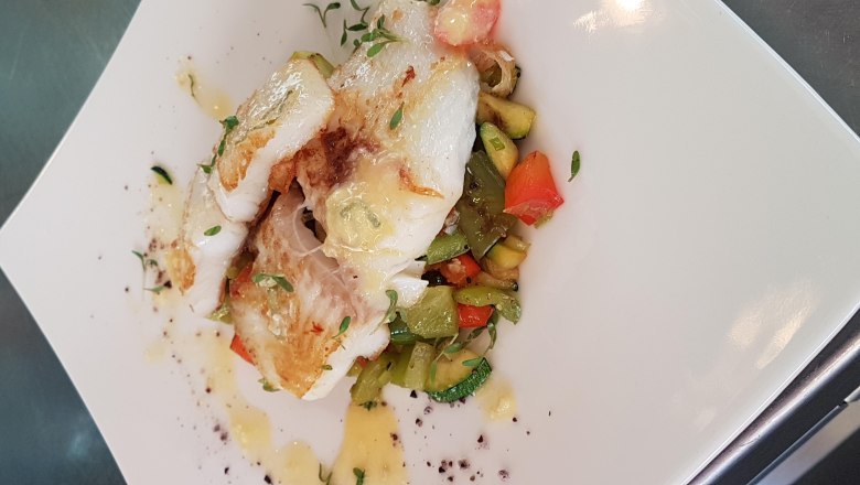 Kabeljau, © Reef & Beef Gebratener Kabeljau auf einem Bett aus buntem Gemüse, serviert auf einem weißen Teller.